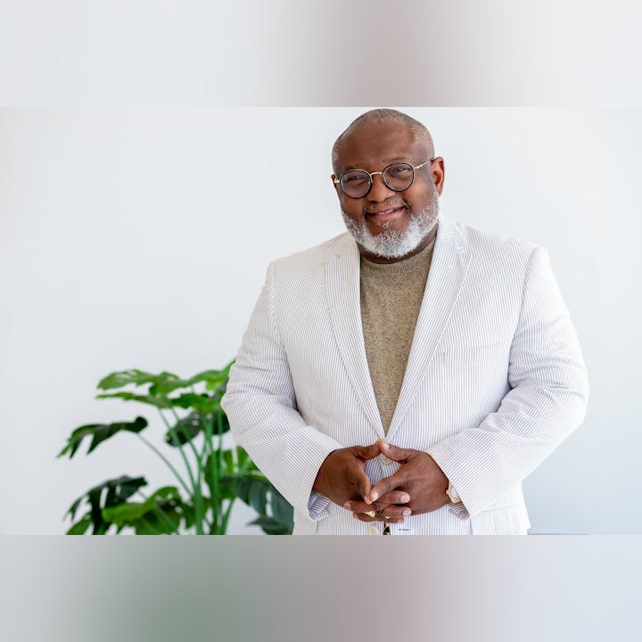 Smiling man in a white striped suit jacket, standing in front of a plant.