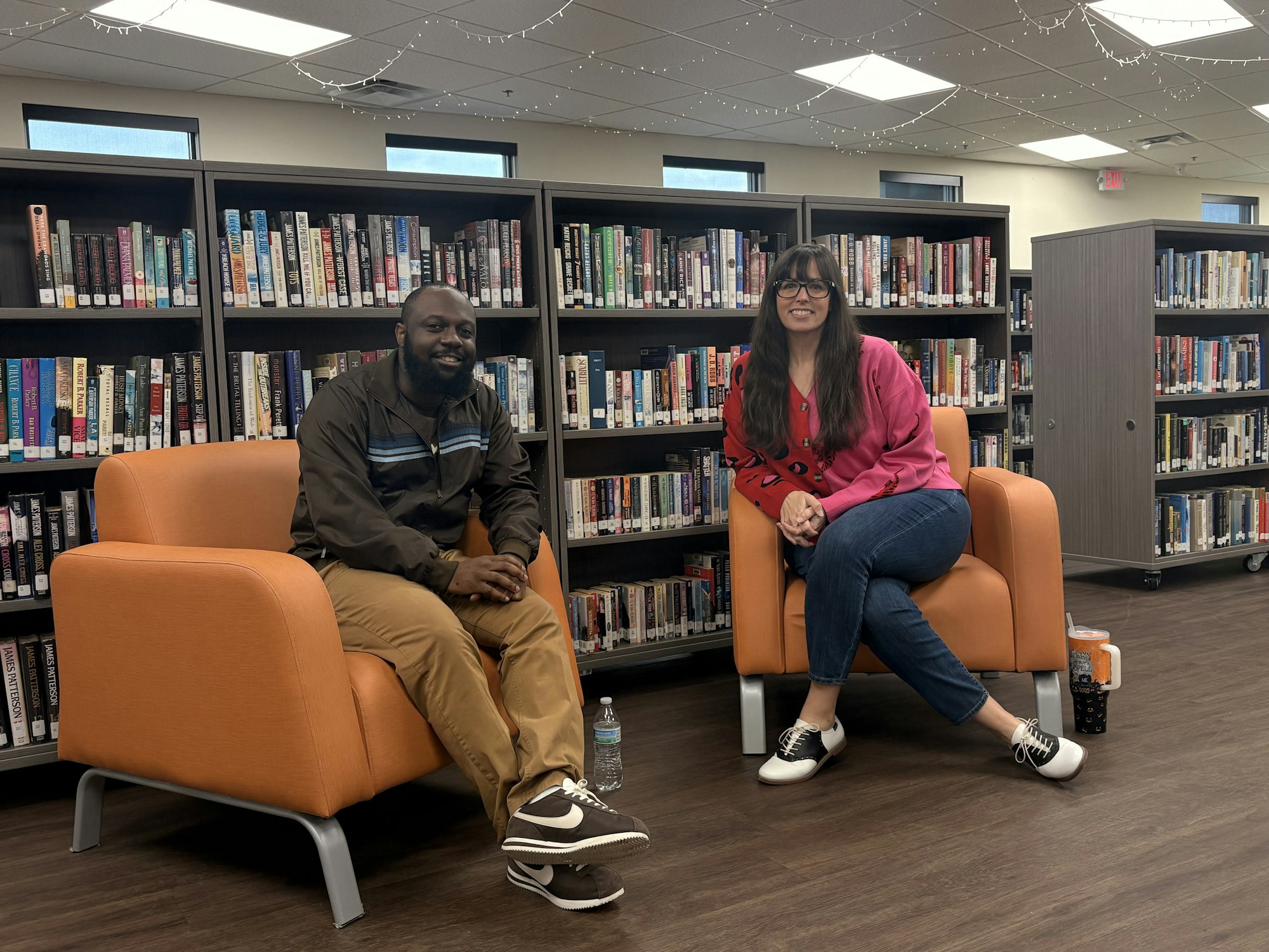 Two individuals sit in orange chairs in a library, surrounded by bookshelves filled with books. Cozy and casual atmosphere.
