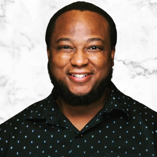 A smiling person with a short beard, wearing a patterned shirt against a marble-like background.