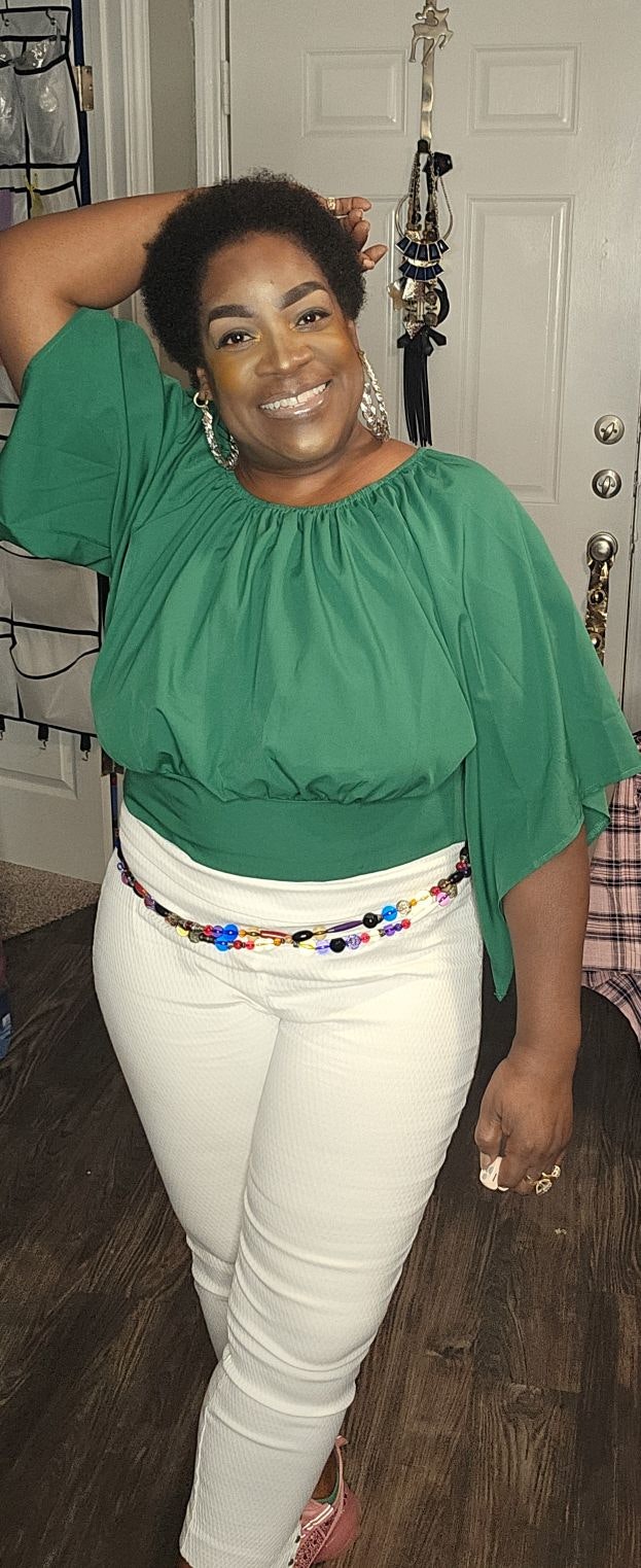 A smiling person in a green blouse and white pants, wearing colorful accessories, posing indoors.