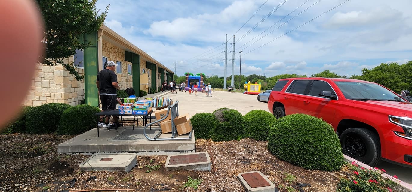 Outdoor event with tables, supplies, inflatable bounce houses, people mingling, a red car, and greenery.