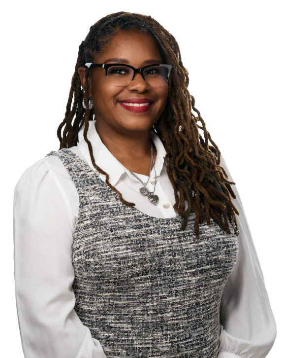 A smiling woman with long, dark, curly hair, wearing glasses and a patterned outfit with a white blouse.