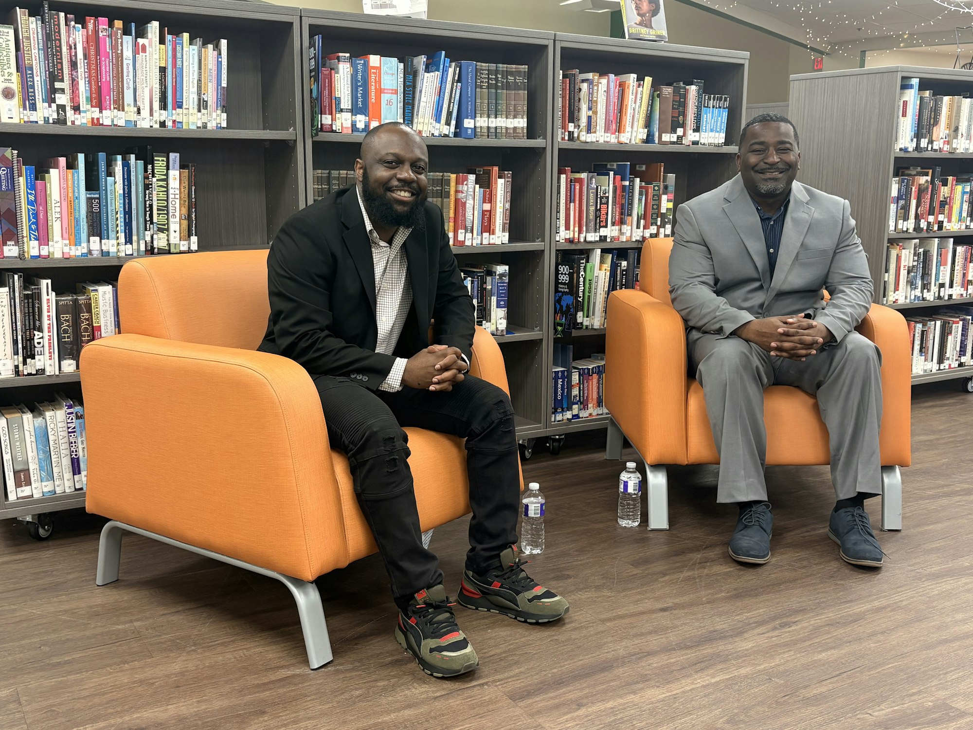 Two men are sitting in orange chairs in a library, surrounded by shelves of books. They appear to be engaged or preparing for an event.