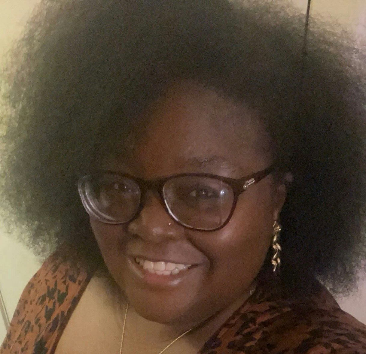A person with glasses, smiling, wearing a patterned top and earrings.
