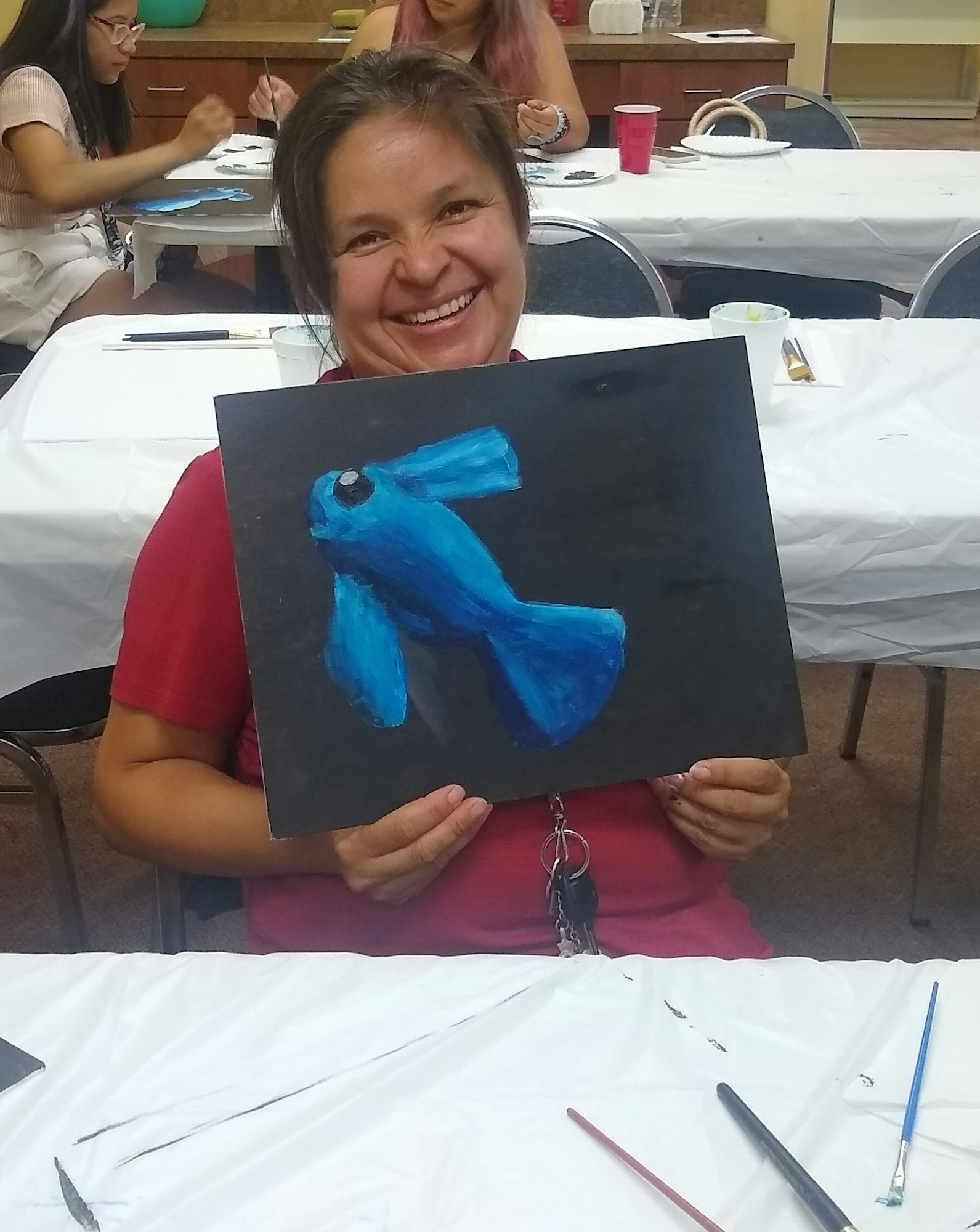 A person smiles while holding up a painting of a blue fish in an art class setting.