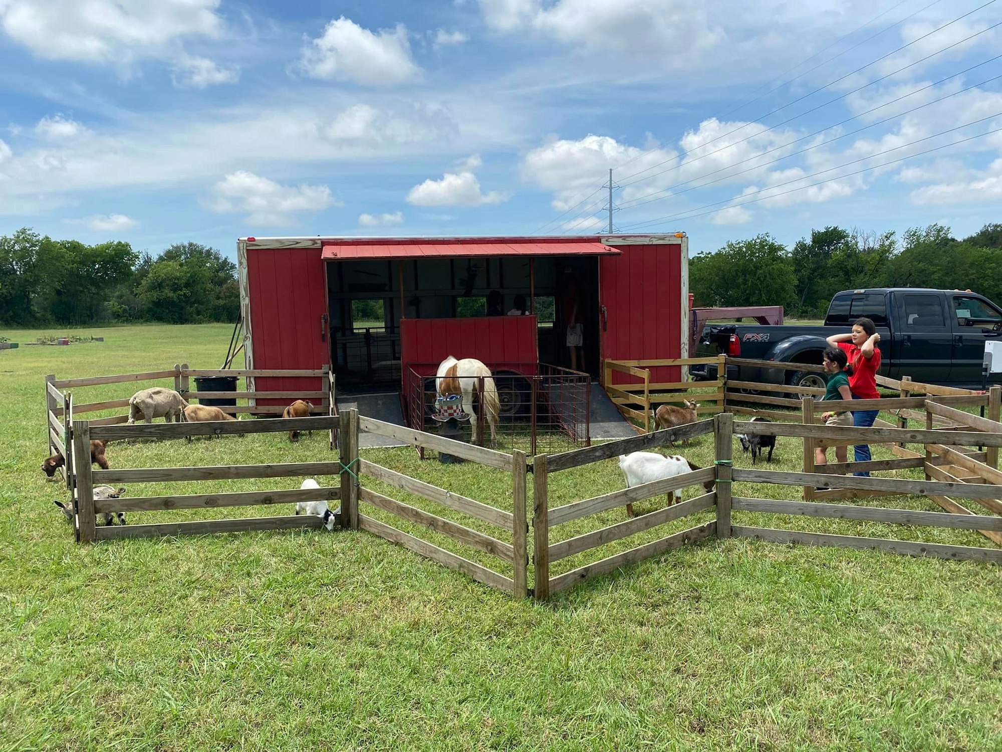 A small red barn with animals inside fenced enclosures, two people observing, and a truck nearby, under a partly cloudy sky.