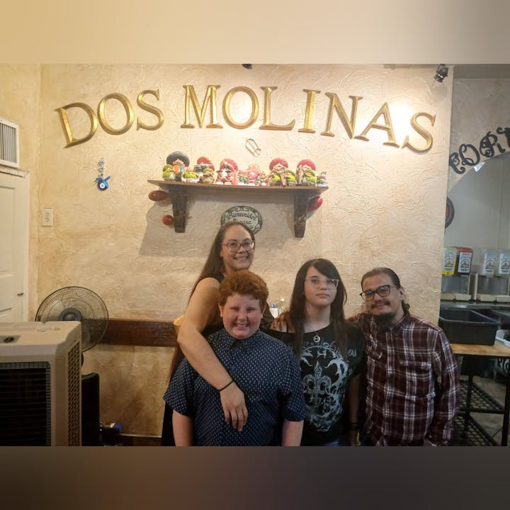 A group of four people stands in front of a restaurant sign "DOS MOLINAS," with decor on the wall behind them.