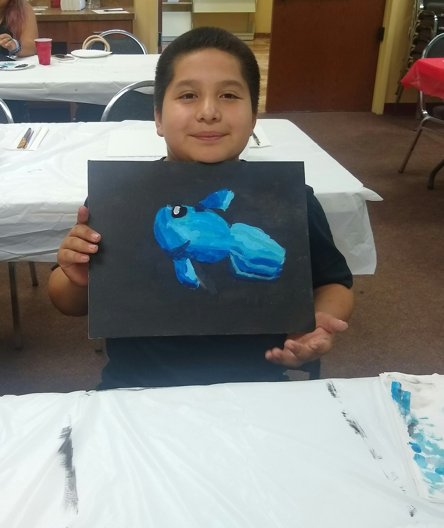 Child holds a painting of a blue fish, smiling, in a classroom with art supplies on tables.