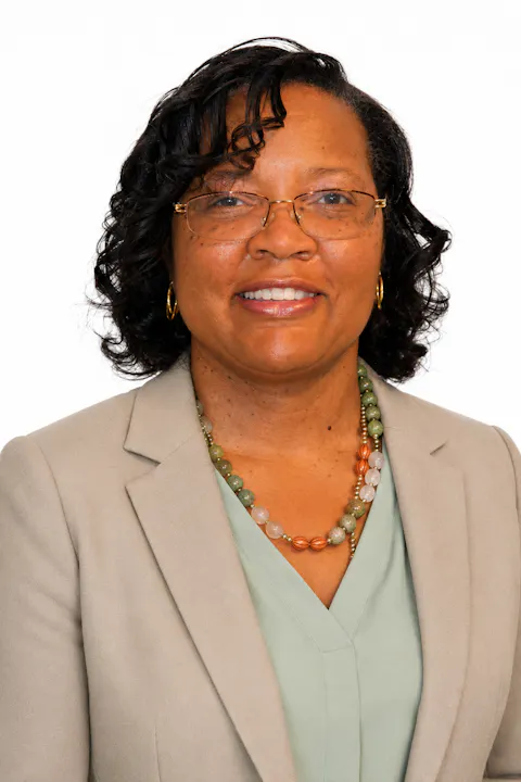 The image shows a woman with curly hair, wearing glasses and a light-colored blazer, smiling against a plain background.