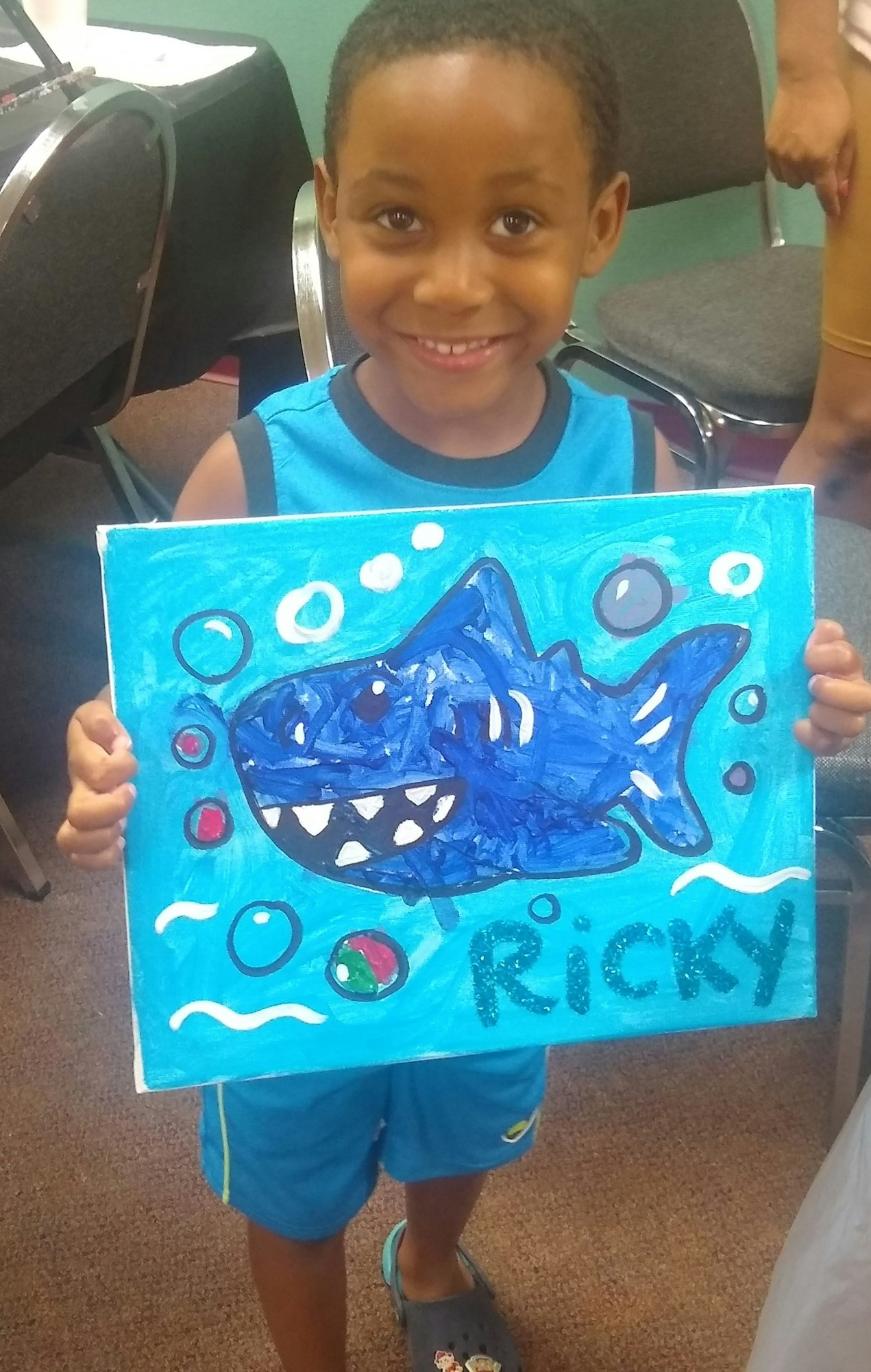 A smiling child holding a bright blue painting of a shark with bubbles, signed "Ricky."