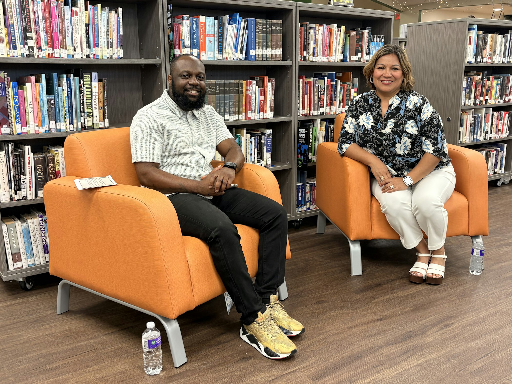 Two individuals sit in orange chairs in a library, surrounded by bookshelves, engaged in a discussion or interview.
