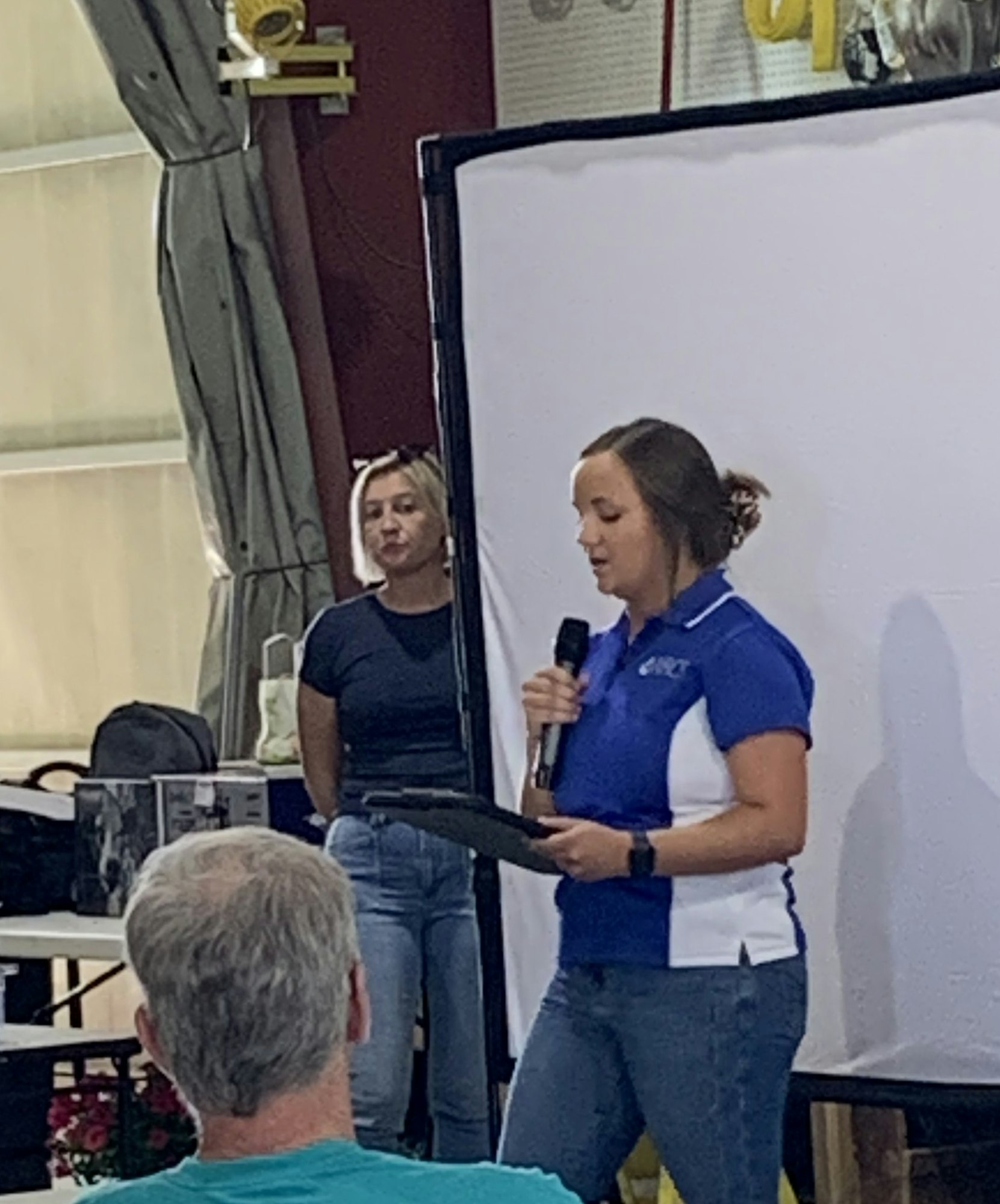 A woman speaks into a microphone in front of a screen, while another woman stands nearby. A man sits in the foreground.