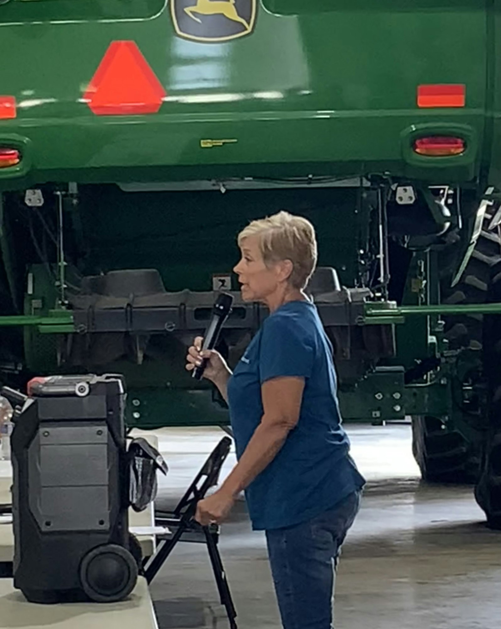 A person speaking into a microphone standing near a green tractor in a indoor setting.