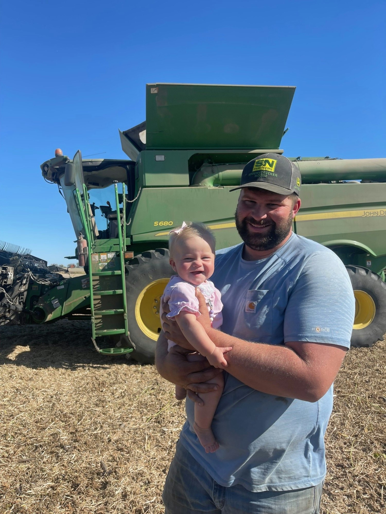 A smiling man holding a baby with a combine harvester in the background.