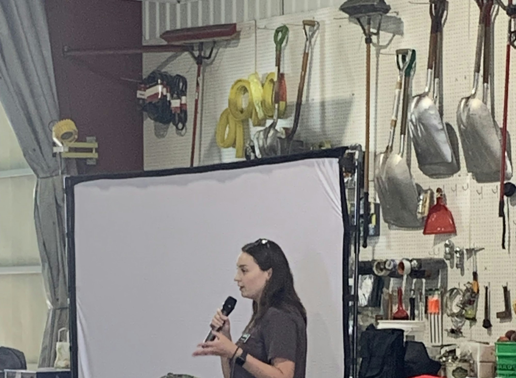 A person holds a microphone speaking in front of a white screen, with tools and gardening equipment on the wall behind.