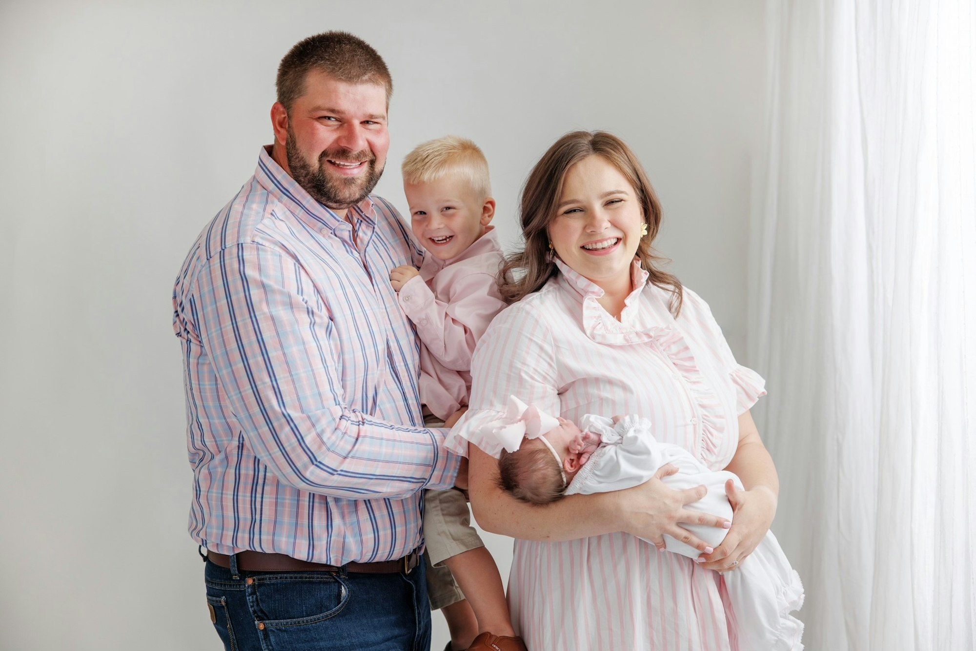 A happy family portrait with a couple, a toddler, and a baby against a white background, all smiling and dressed in pink and white.