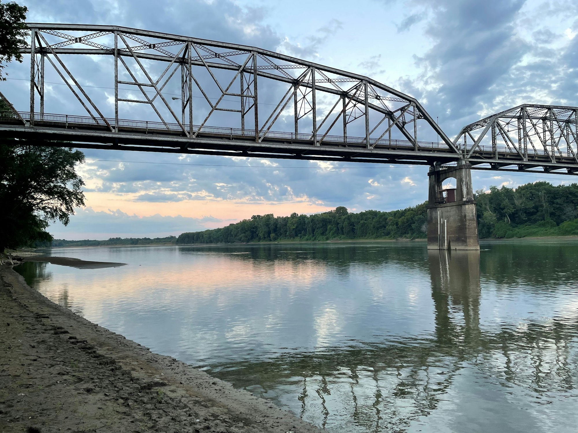 A metal bridge over a calm river with a sandy shore and trees, under a cloudy sky.