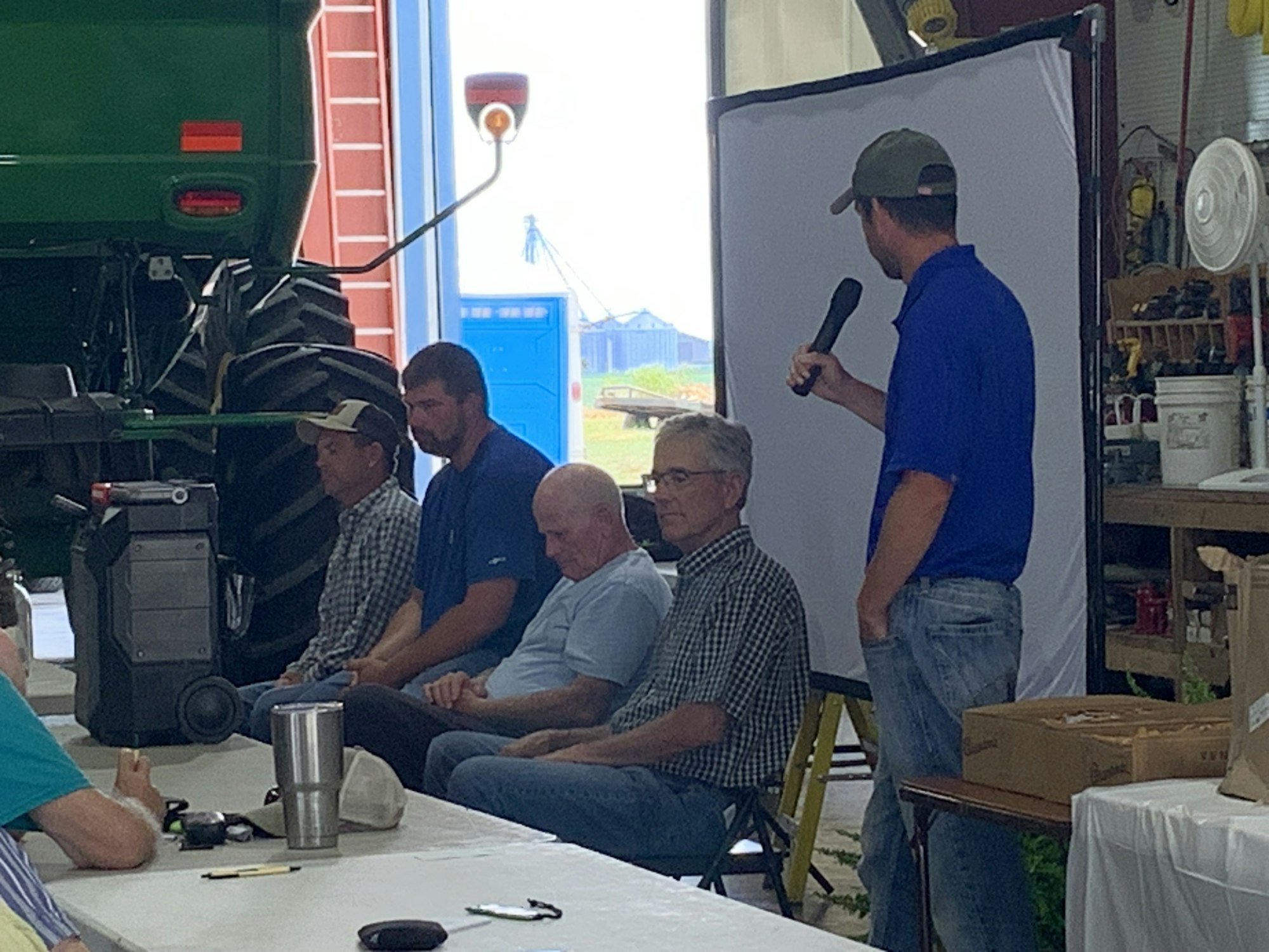 Four men sit, one stands talking into a microphone near a projector screen. Machinery and tools are visible.