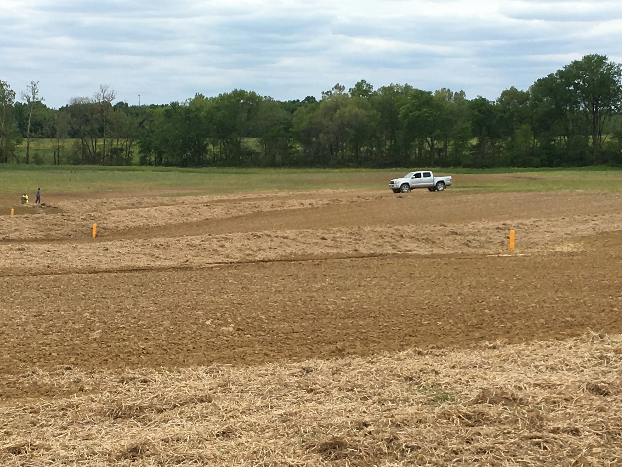 A truck on plowed farmland with two people and survey markers. Trees in the background under a cloudy sky.