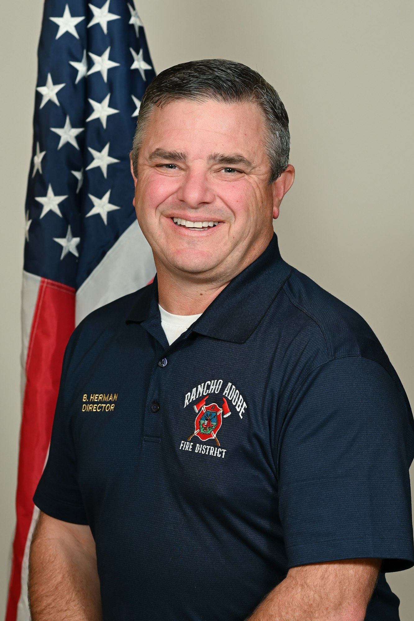 A person in a uniform with "Rancho Adobe Fire District" logo, standing in front of a U.S. flag.