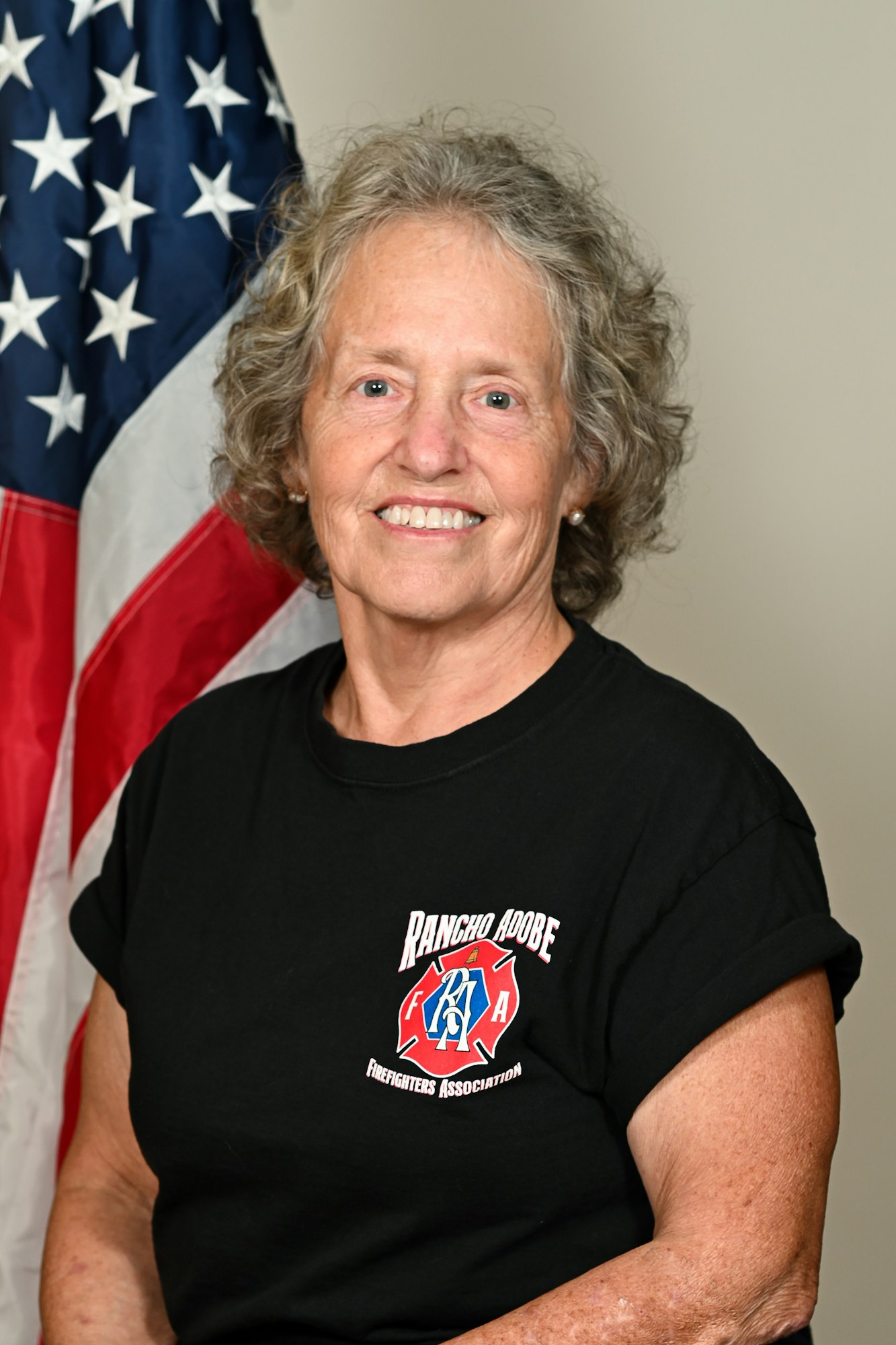 A person in a black "Rancho Adobe Firefighters Association" shirt poses in front of a U.S. flag.