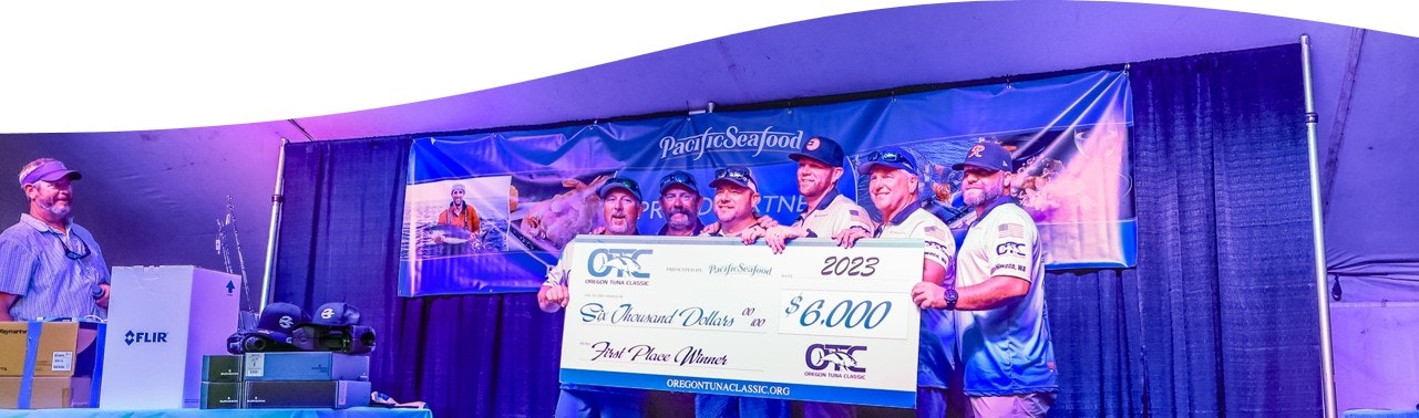 Group of people holding a large check for $6,000 at a fishing event, with branded banners and boxes in the background.