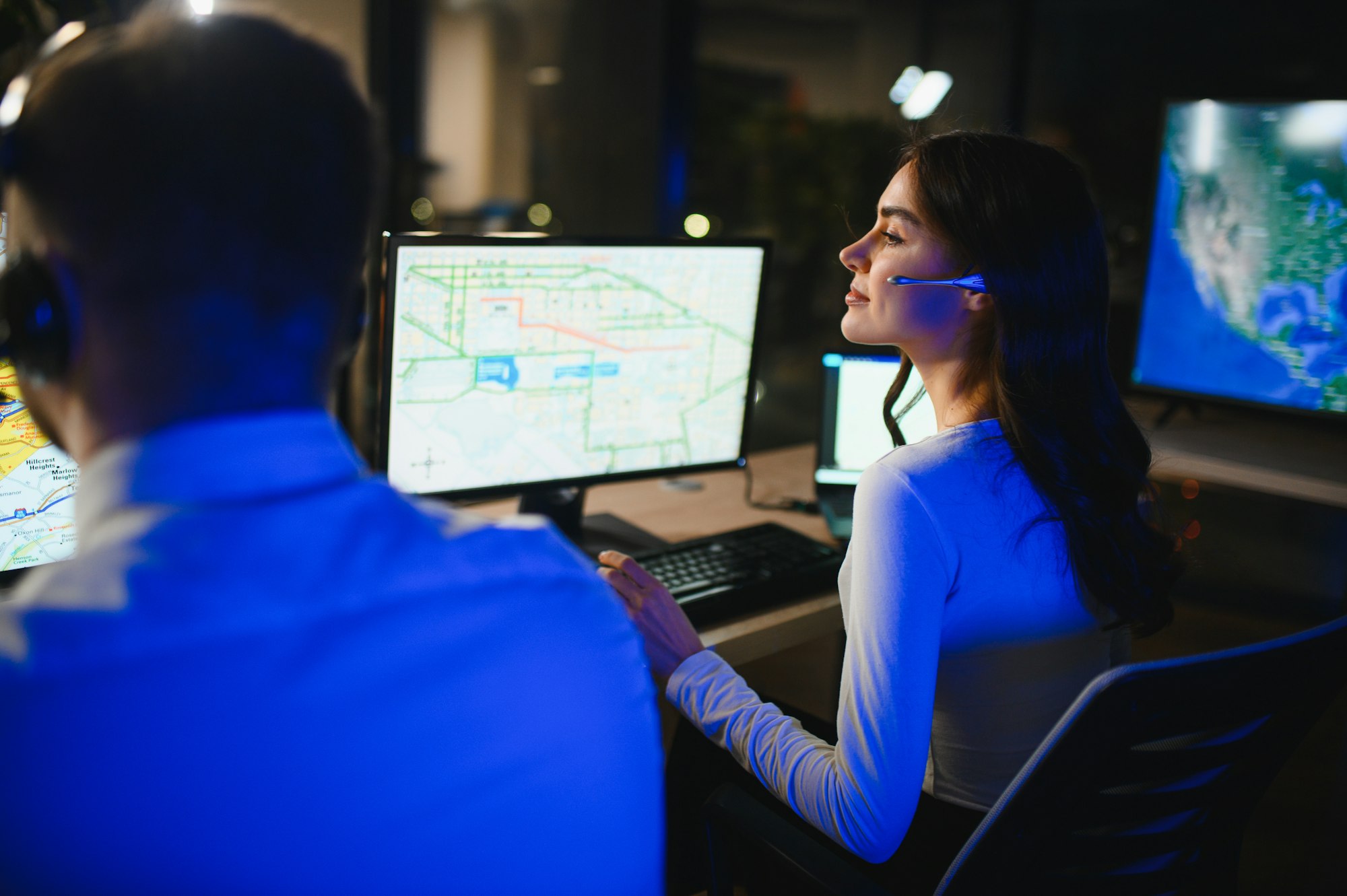 A woman with a headset is focused on multiple computer screens displaying maps in a dark office setting.