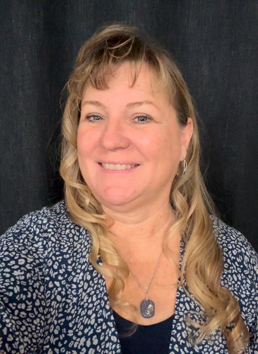 A person with long hair is smiling at the camera, wearing a patterned top and a necklace, against a dark background.