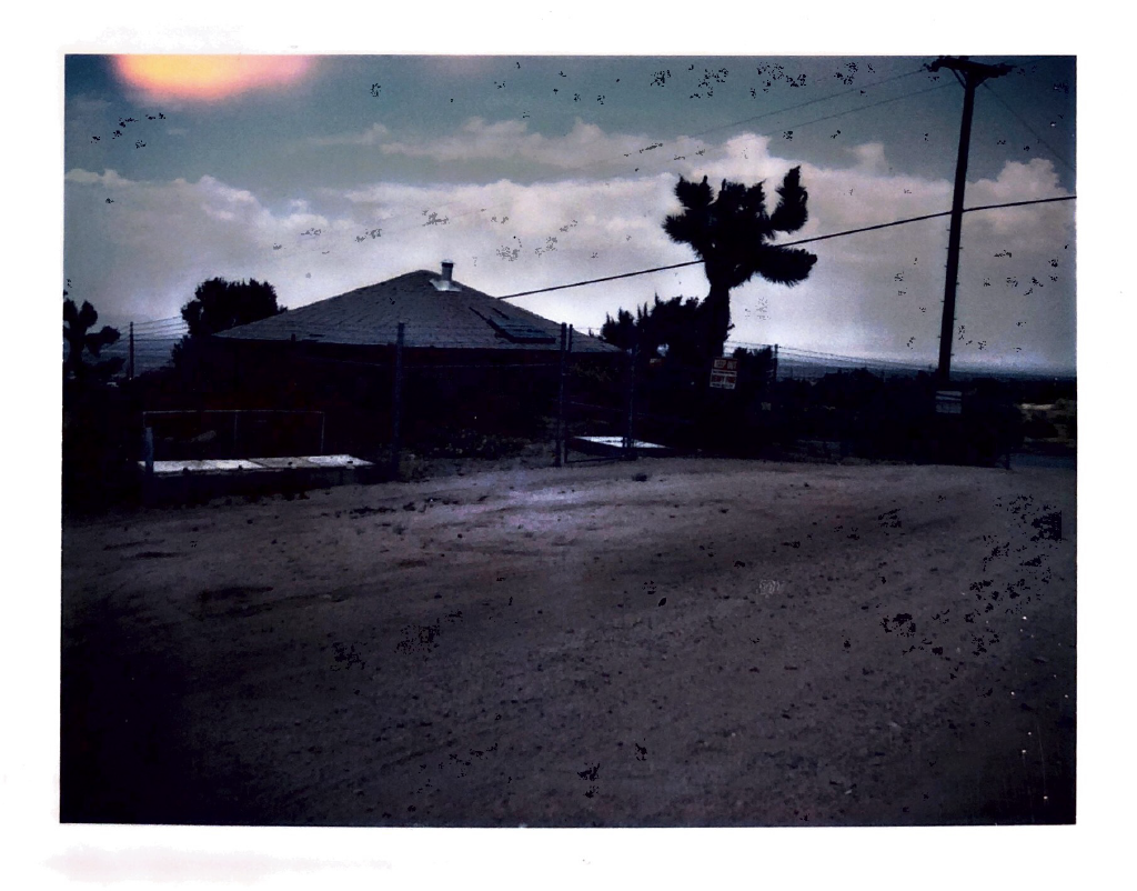 A weathered house near a dirt road, surrounded by trees and power lines, under a cloudy sky.