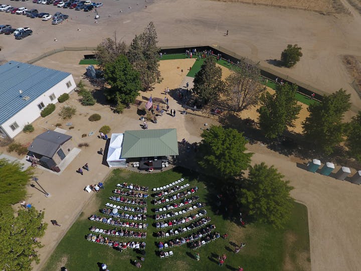 An aerial view of an outdoor event with seated attendees, a stage and tents, surrounded by trees and parking.