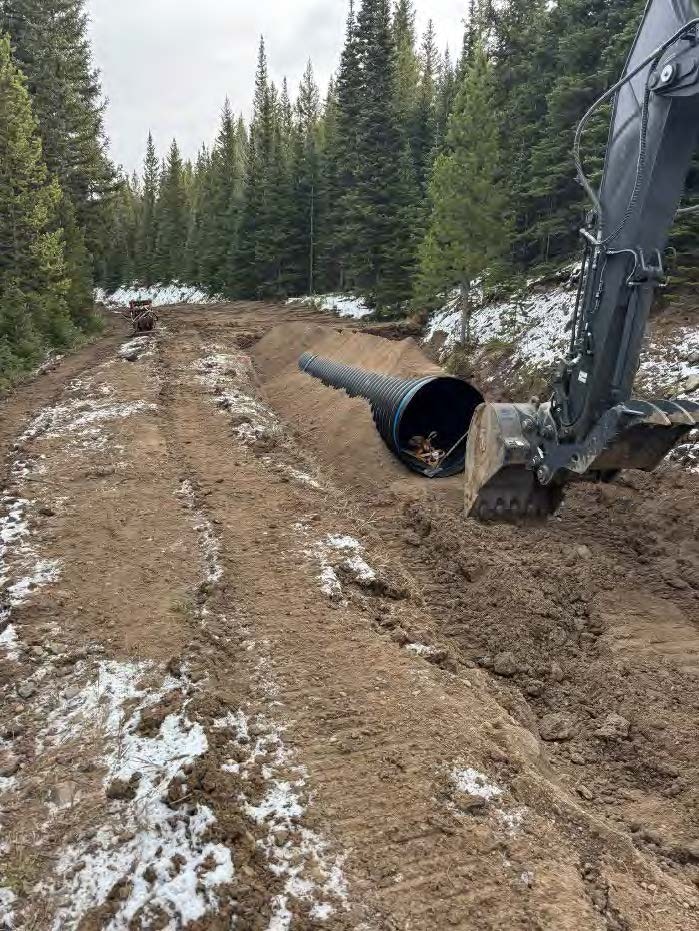 The image shows a construction site with a large black pipe on a dirt road, surrounded by trees and some snow.