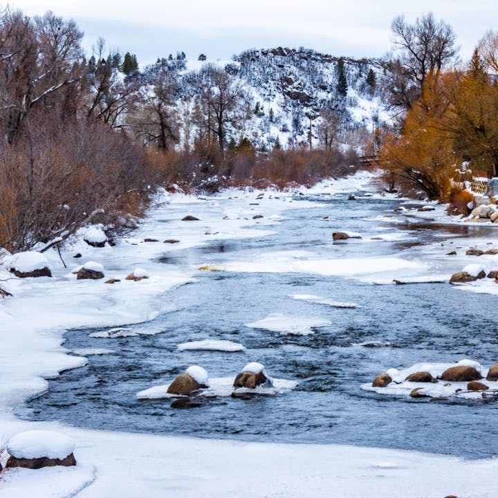 A snowy river scene, with rocks and snowy banks, surrounded by trees and hills in the background.