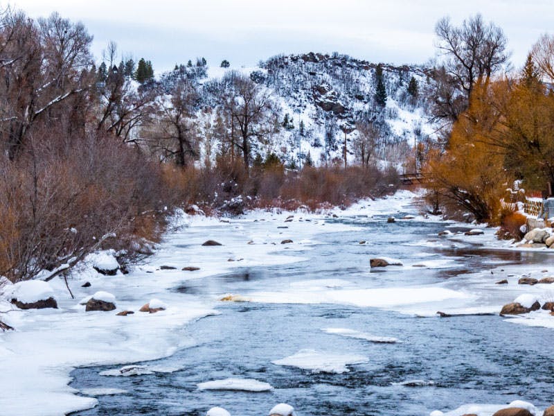 A snowy river scene, with rocks and snowy banks, surrounded by trees and hills in the background.