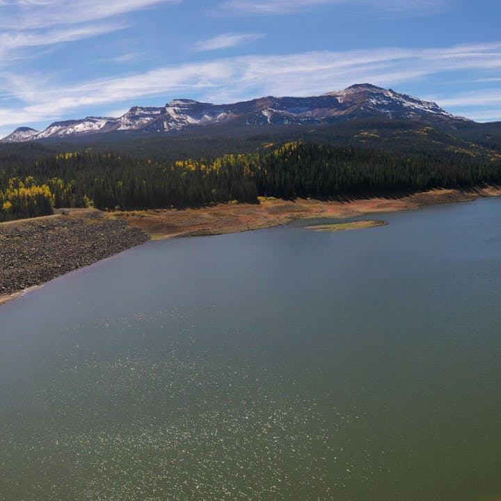A serene lake with a forested shoreline and snow-capped mountains in the background under a clear blue sky.