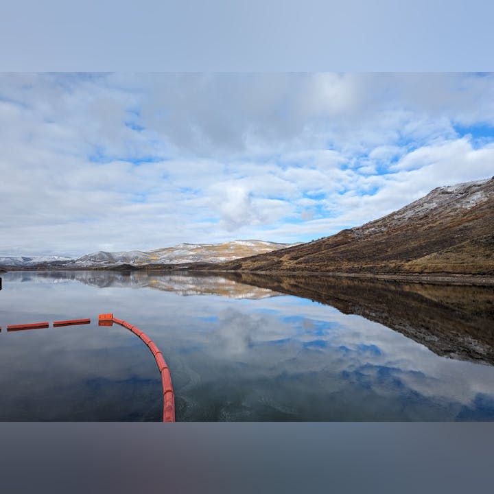 A calm lake reflecting clouds and mountains, with a red barrier in the foreground under a partly cloudy sky.
