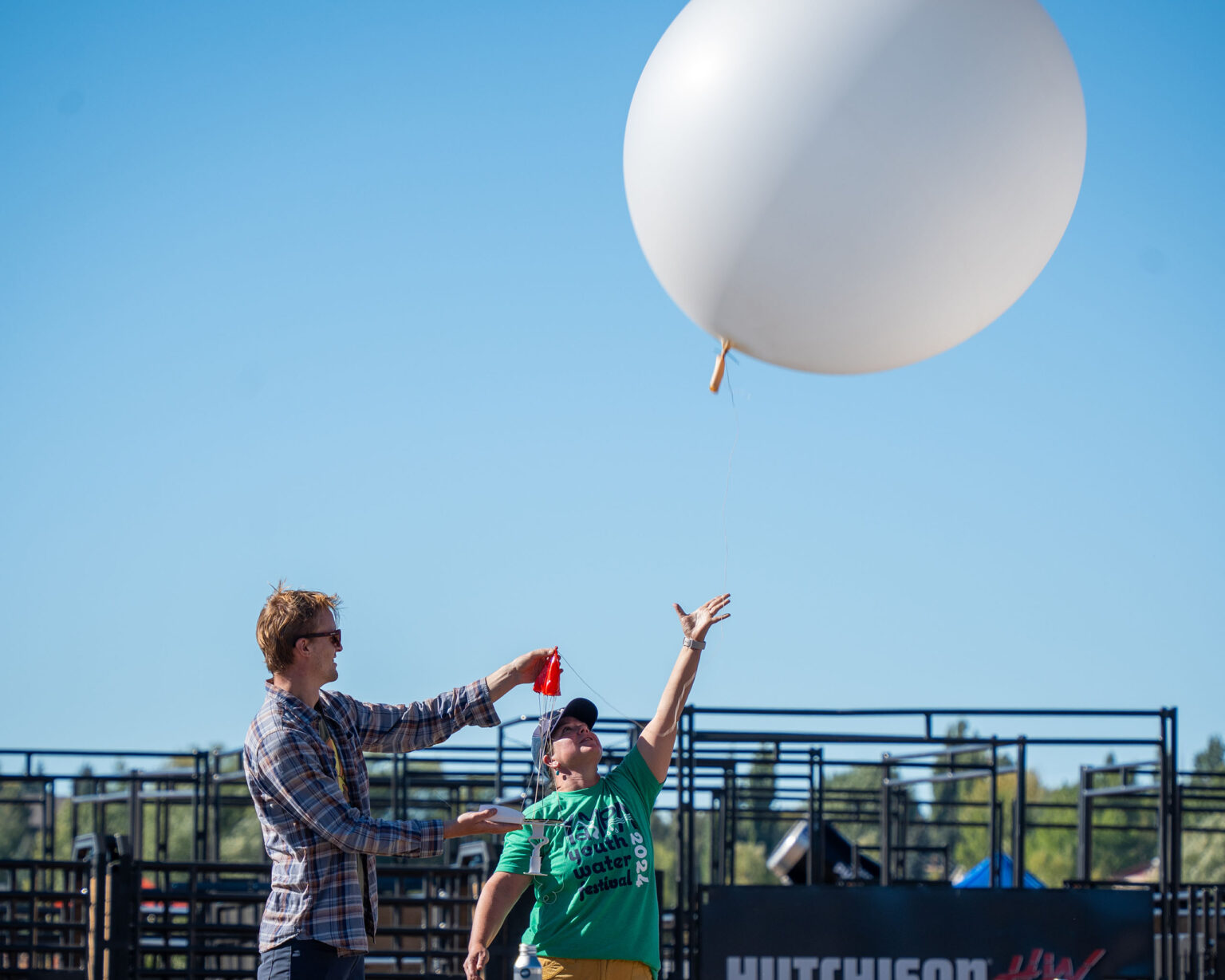 Two people outdoors, one holding a large balloon, the other releasing a red cup, with blue sky and metal structure in the background.