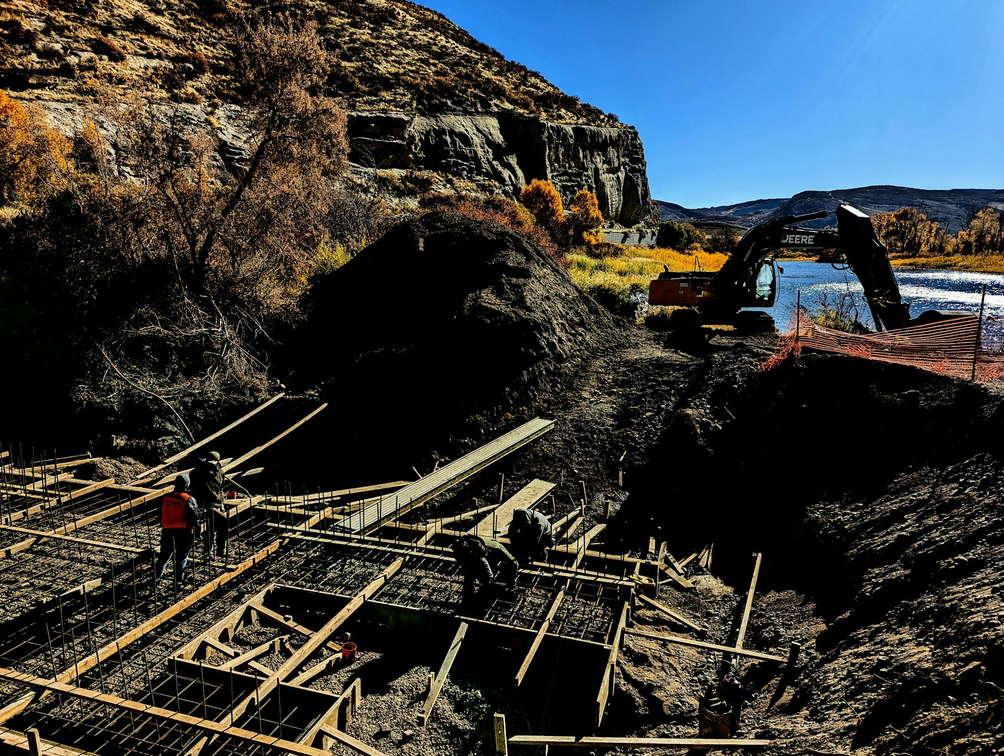 Construction site with workers, framing, and an excavator near a river and hills under a clear blue sky.