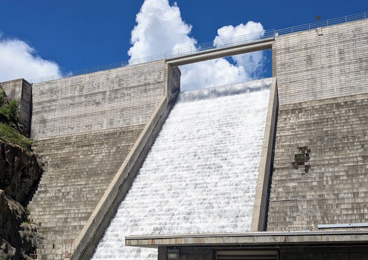 A dam spillway with water flowing down, set against a bright blue sky with clouds overhead.