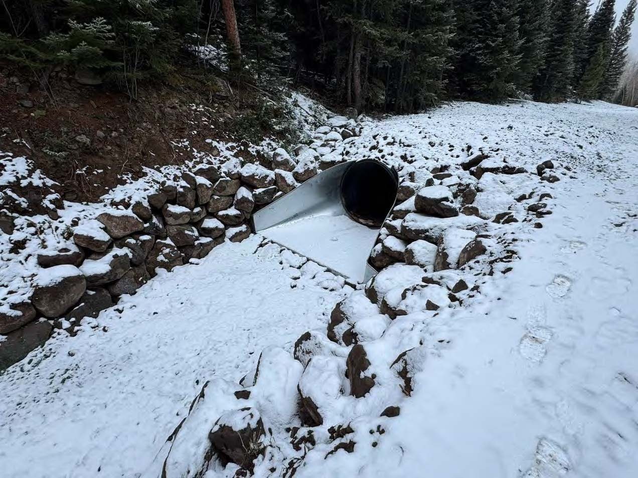 A snow-covered ground with a metallic pipe surrounded by rocks in a wooded area. Footprints are visible in the snow.
