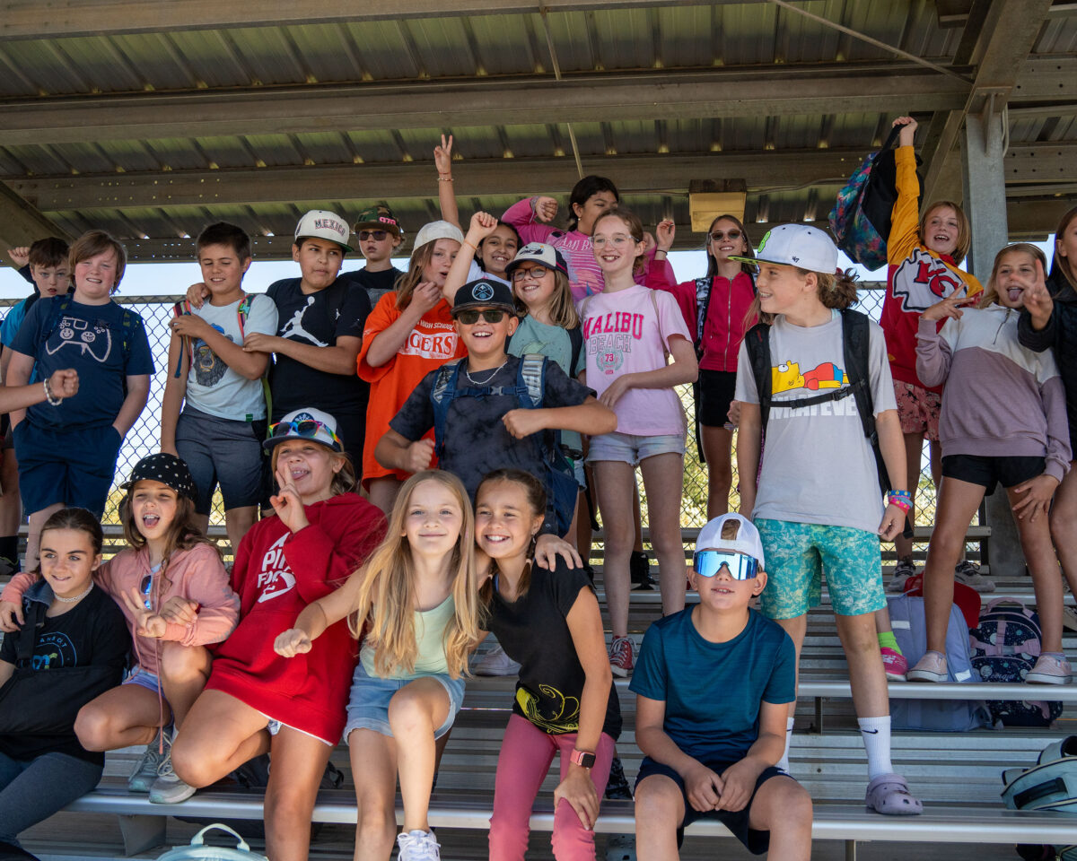 A group of kids posing cheerfully on bleachers, some wearing caps and sunglasses, with backpacks in the background.