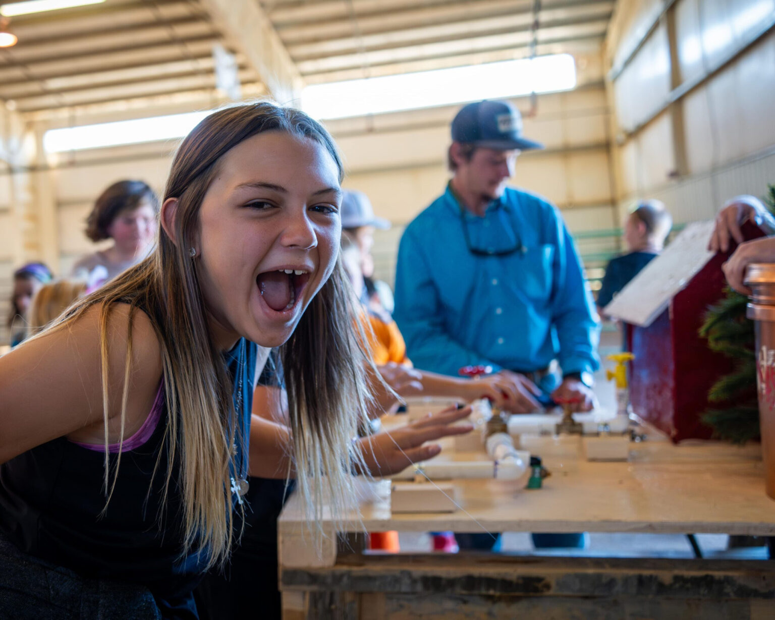 A girl excitedly poses in front of people working on a hands-on project at a workshop or event.