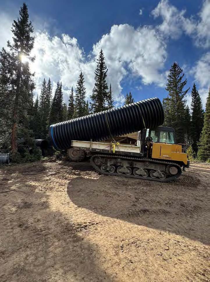 A tracked vehicle is moving large black pipes in a forested area under a partly cloudy sky.