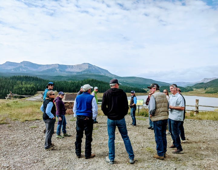 A group of people outdoors near a lake with mountains and forest in the background.