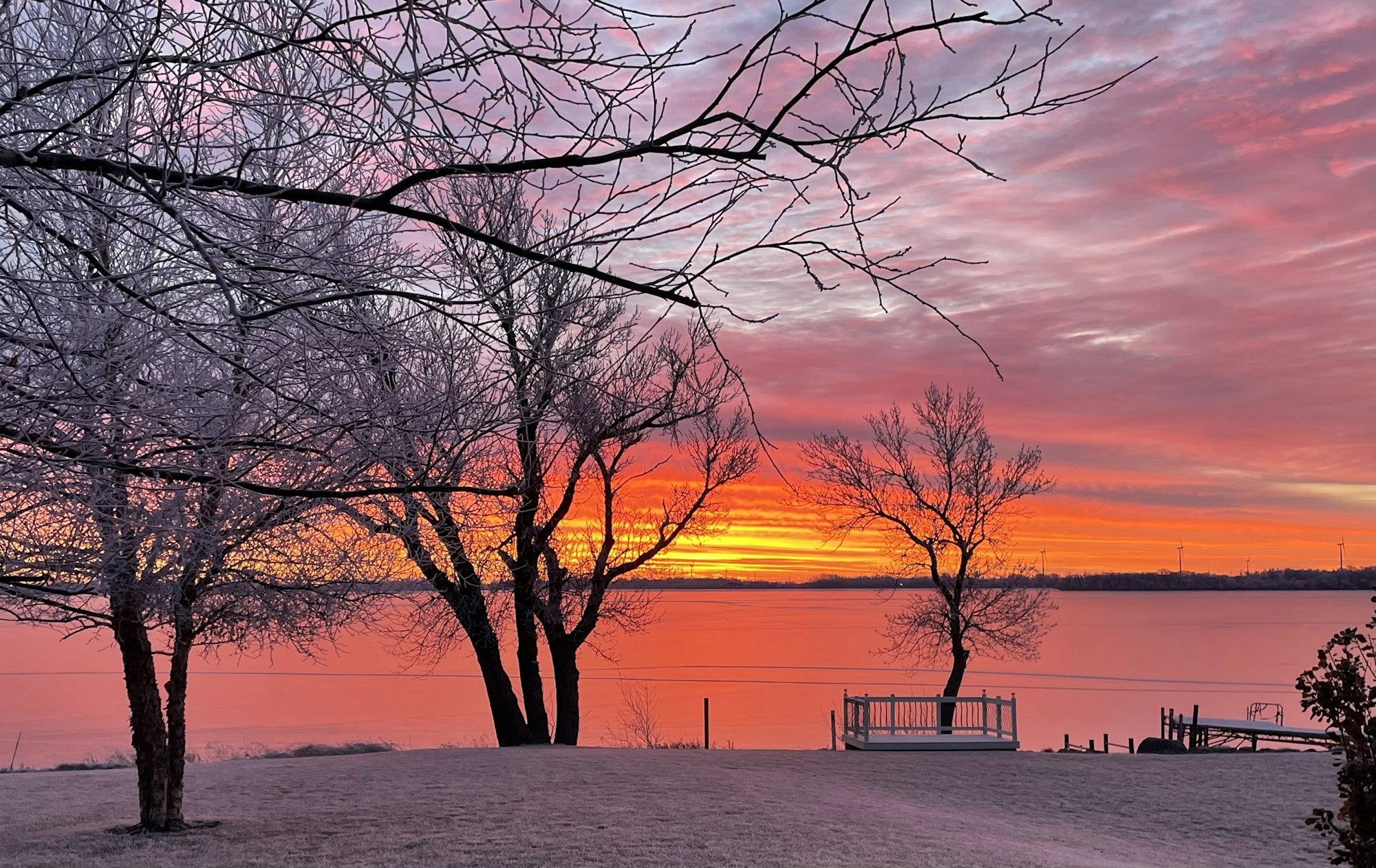 A frosty landscape with bare trees at sunrise over a calm lake, with a vibrant pink and orange sky.