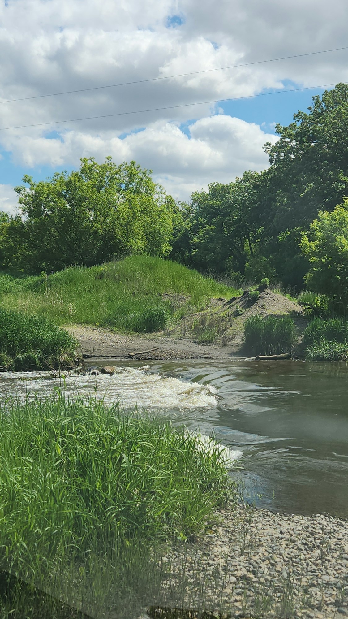 A peaceful river scene with green grass, trees, and a cloudy blue sky.