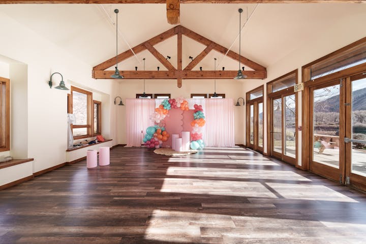 A spacious room with wooden beams, large windows, pink balloons, and a decorative backdrop, suitable for events or gatherings.
