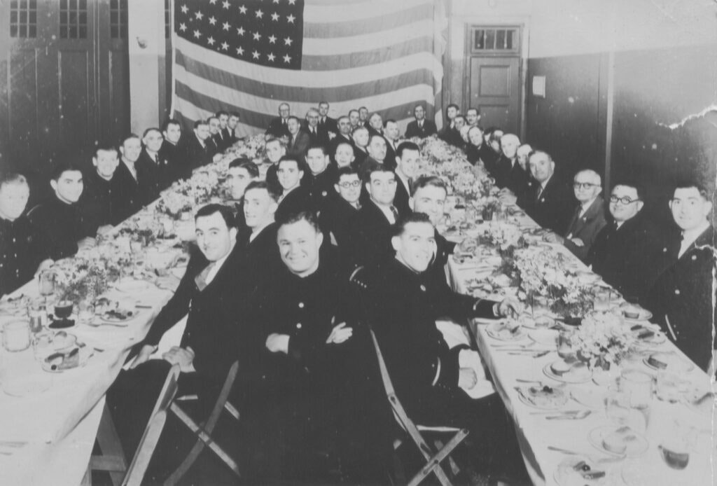 A large group of people dressed formally seated at a long banquet table, with an American flag in the background.