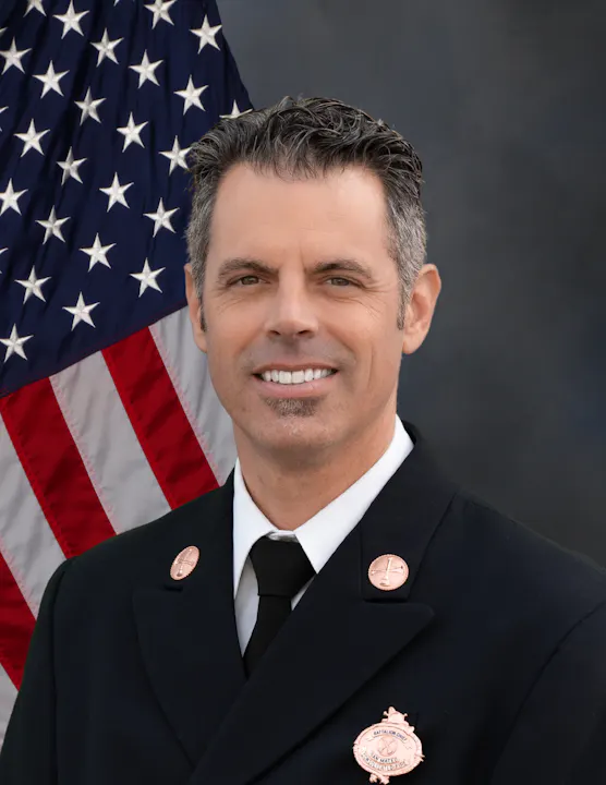 A professional portrait of a man in a fire department uniform, smiling with an American flag background.