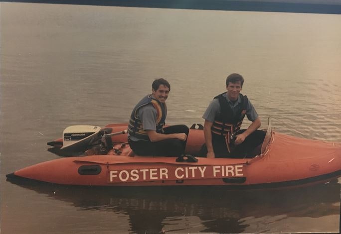 Two people in life vests are seated on a red watercraft labeled "Foster City Fire" on a body of water.