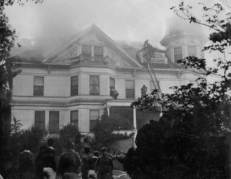 A large house with smoke, people using ladders, and onlookers nearby.